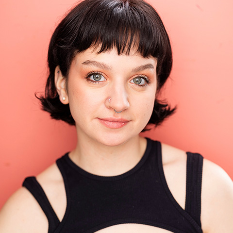Bella Donneruno headshot: grinning in black top in front of peach wall