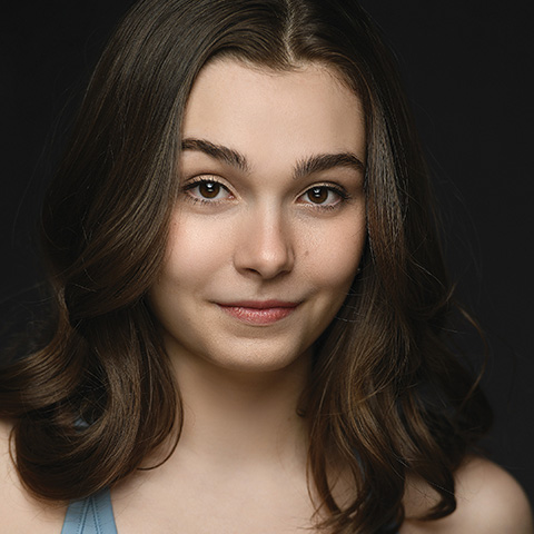 Olivia Foght headshot: grinning in blue tank top in front of black wall