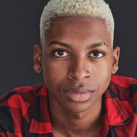 Jordan Hall headshot: wearing red and black plaid shirt in front of black wall
