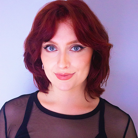 Rebecca Jensen headshot: wearing black sheer top in front of a lilac wall