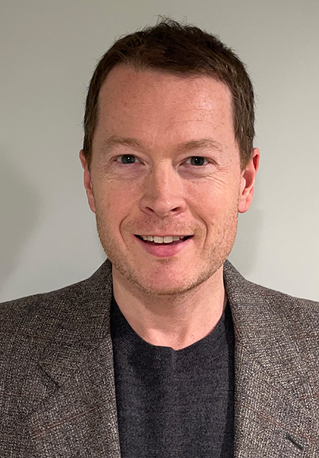 Andrew Shryock headshot; wearing black shirt with gray blazer in front of a white/gray background