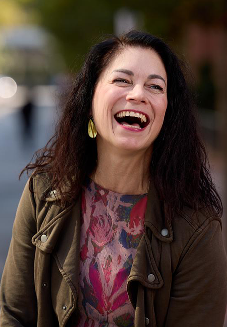Liz Roncka smiling–mid laugh. She is wearing a colorful (red, blue, purple) shirt with a brown jacket.