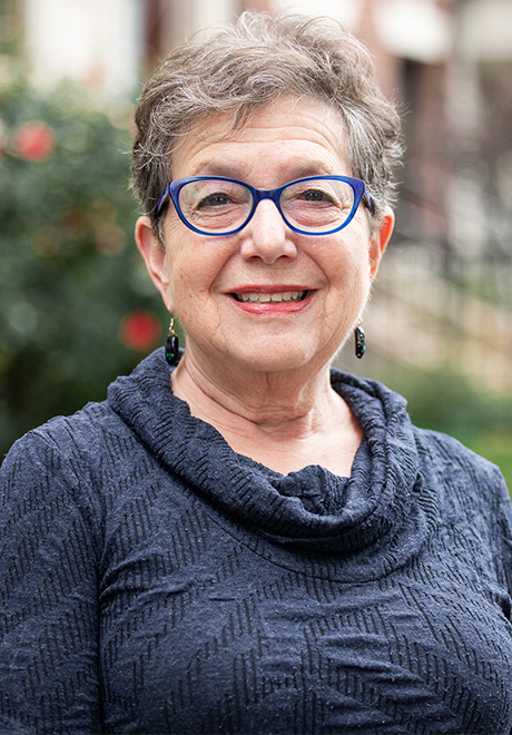 Sara Goldstein headshot: smiling in front of buildings and wearing a blue/gray sweater.