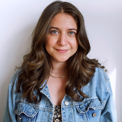 Alexis Scheer headshot: smiling and wear a jean jacket in front of a white wall.