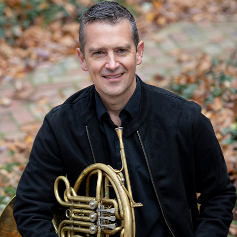 Adam Unsworth headshot: sitting outside with leaves in the background, wearing all black and holding his french horn.