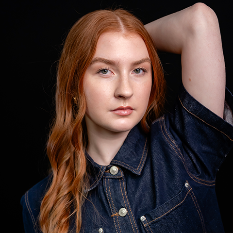 Michaela Harmon headshot: posing in a denim top with right arm angled with right hand on the back of head. Background is black.
