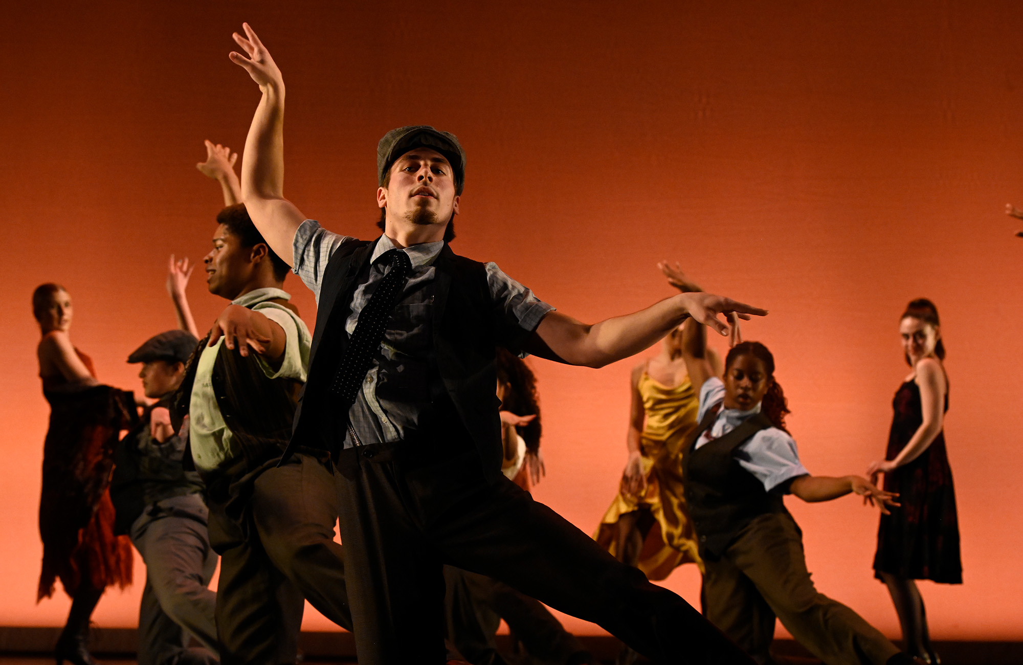 Dancers on stage in front of a n ornage background. Dancer in foreground has left hand lifted towards the ceiling with the right hands horizontal towards the right side.