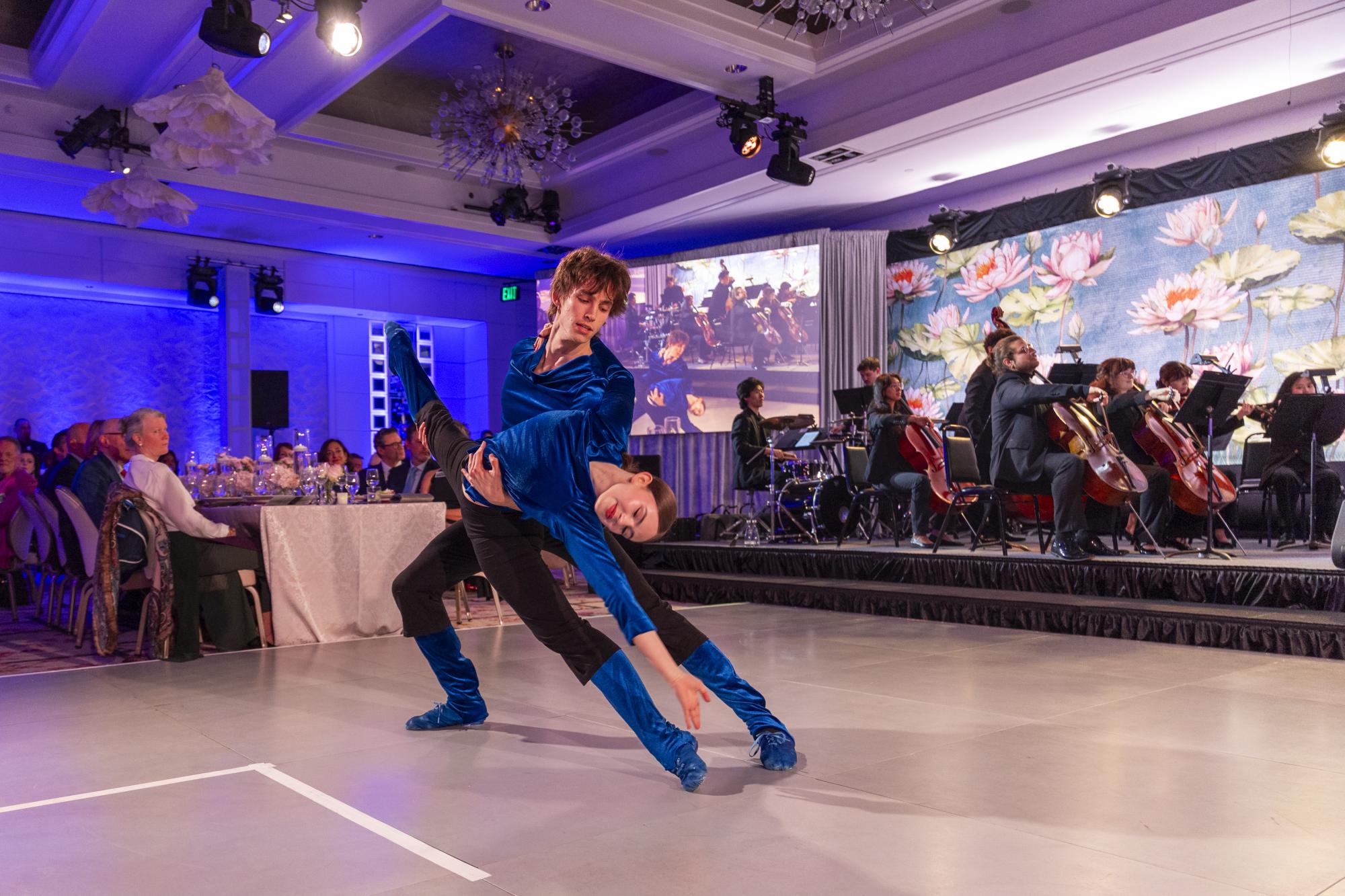 two dance students performing at the Soirée in black and black outfits. One student is holding the other's leg up to the sky.