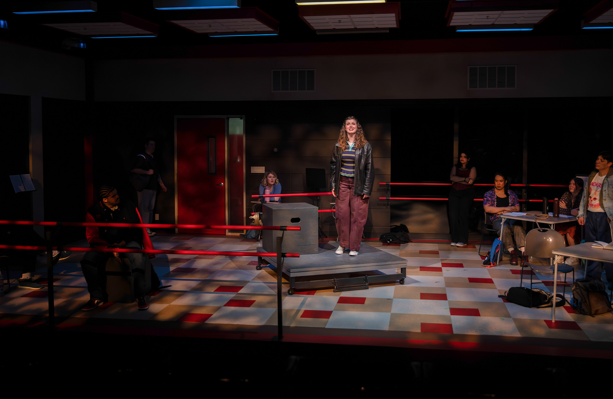 person singing on stage with a spotlight and small podium beneath their feet. The floor is checkered.