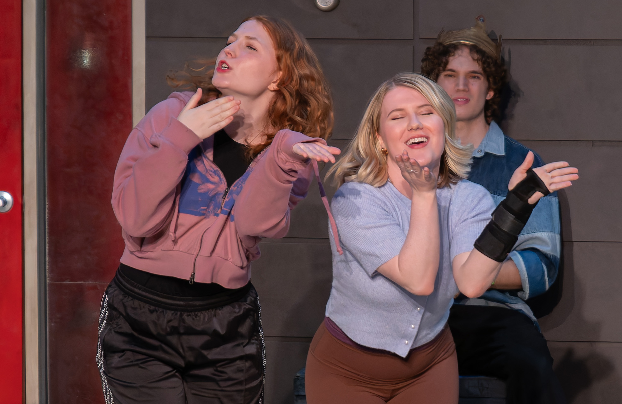 Students performing in 'Time to Act' laughing and singing onstage. One girl is wearing a pink hoodie and track pants, and another is wearing a light blue cardigan. A man in the back in wearing a blue shirt.