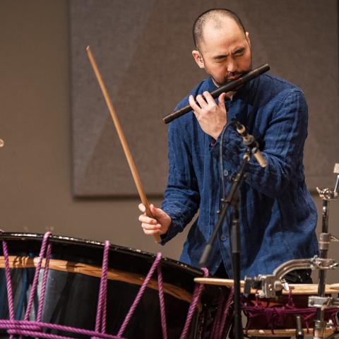 Performer playing a wooden flute with one hand and hitting a drum in the other hand