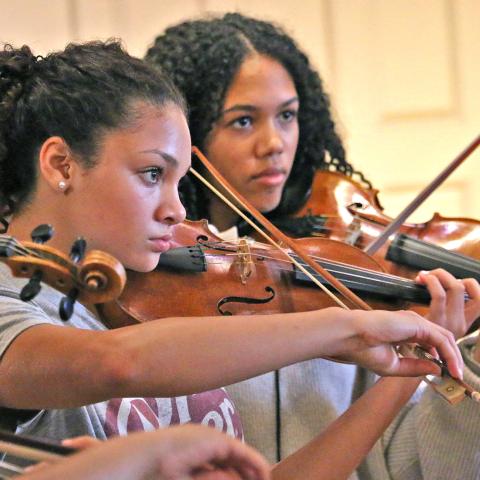 Two young violinists from Sphinx Performance Academy rehearse in Seully Hall. Both are wearing gray shirts holding their instrument up and playing.