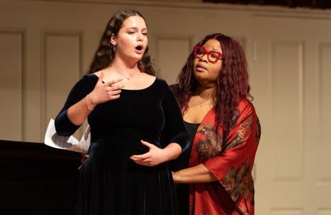 Jamila Drecker-Waxman (left, in black dress, singing) and Karen Slack (right, in red dress) in a performance hall.