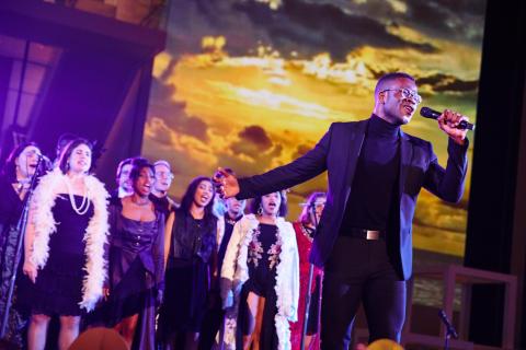 Person singing in front of a choir with clouds in the background 