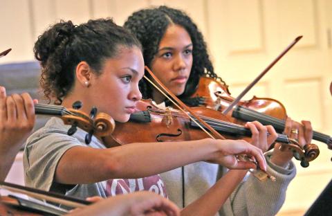 Two young violinists from Sphinx Performance Academy rehearse in Seully Hall. Both are wearing gray shirts holding their instrument up and playing.